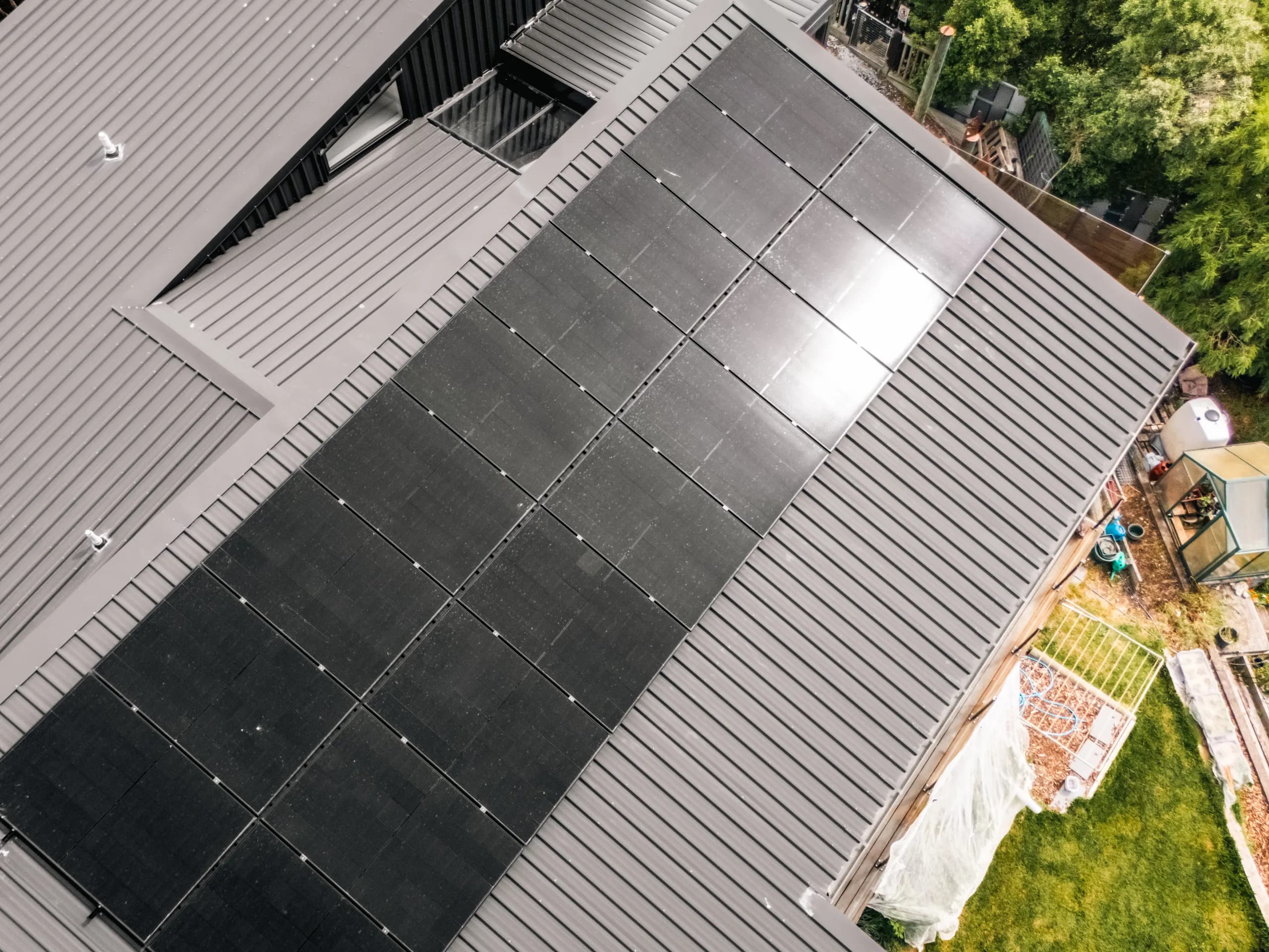 Top-down drone view of a black solar array installed on a dark residential metal roof with a backyard garden below.