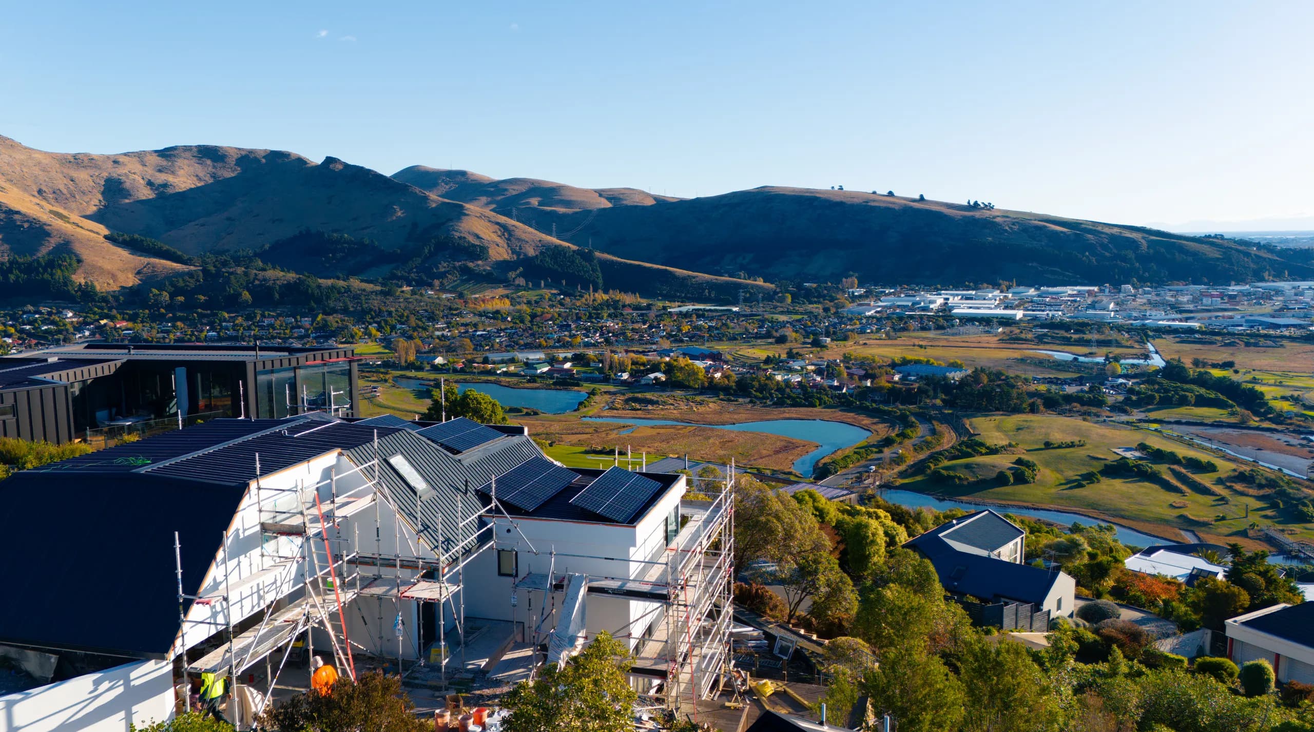 Aerial view of a scaffolded hillside home with rooftop solar panels overlooking a Canterbury valley, wetlands and township at golden hour.