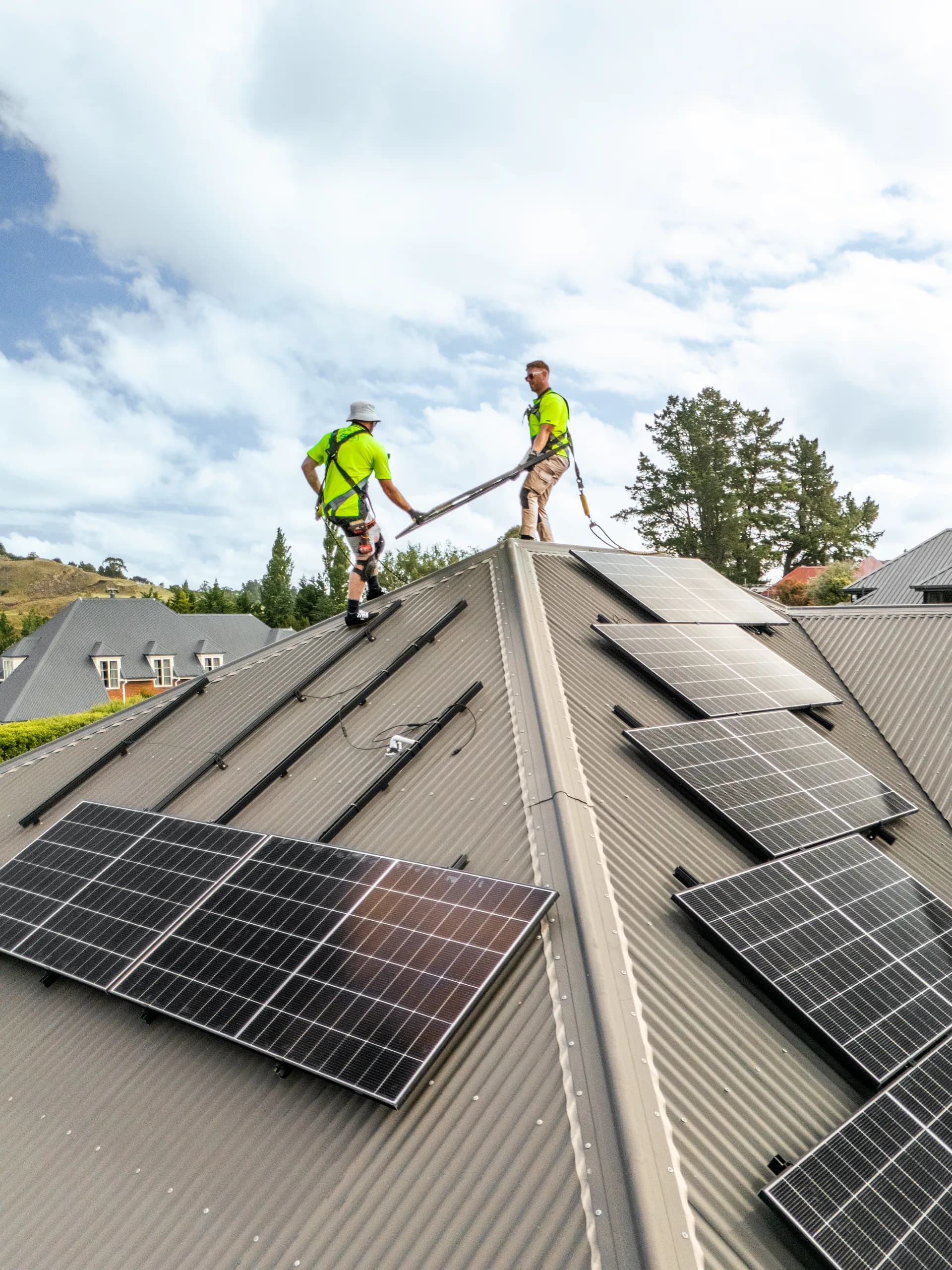 Two ZERO electricians in harnesses fit solar panels to a dark corrugated house roof in a residential neighbourhood.
